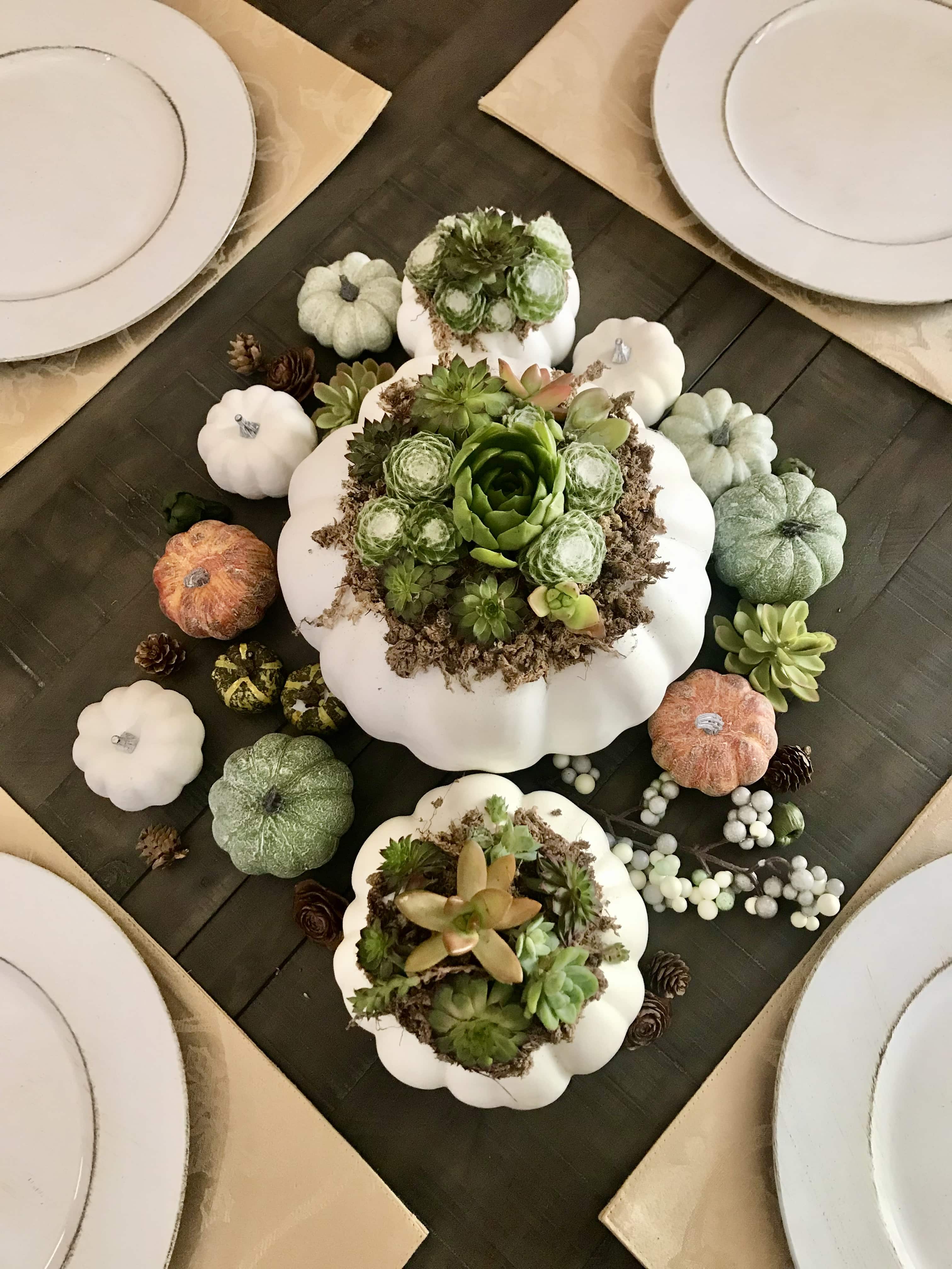 A Succulent pumpkin table centerpiece. Large white pumpkin with succulents surrounded by smaller faux pumpkins at the center of the table. Placemats and white plates also shown.