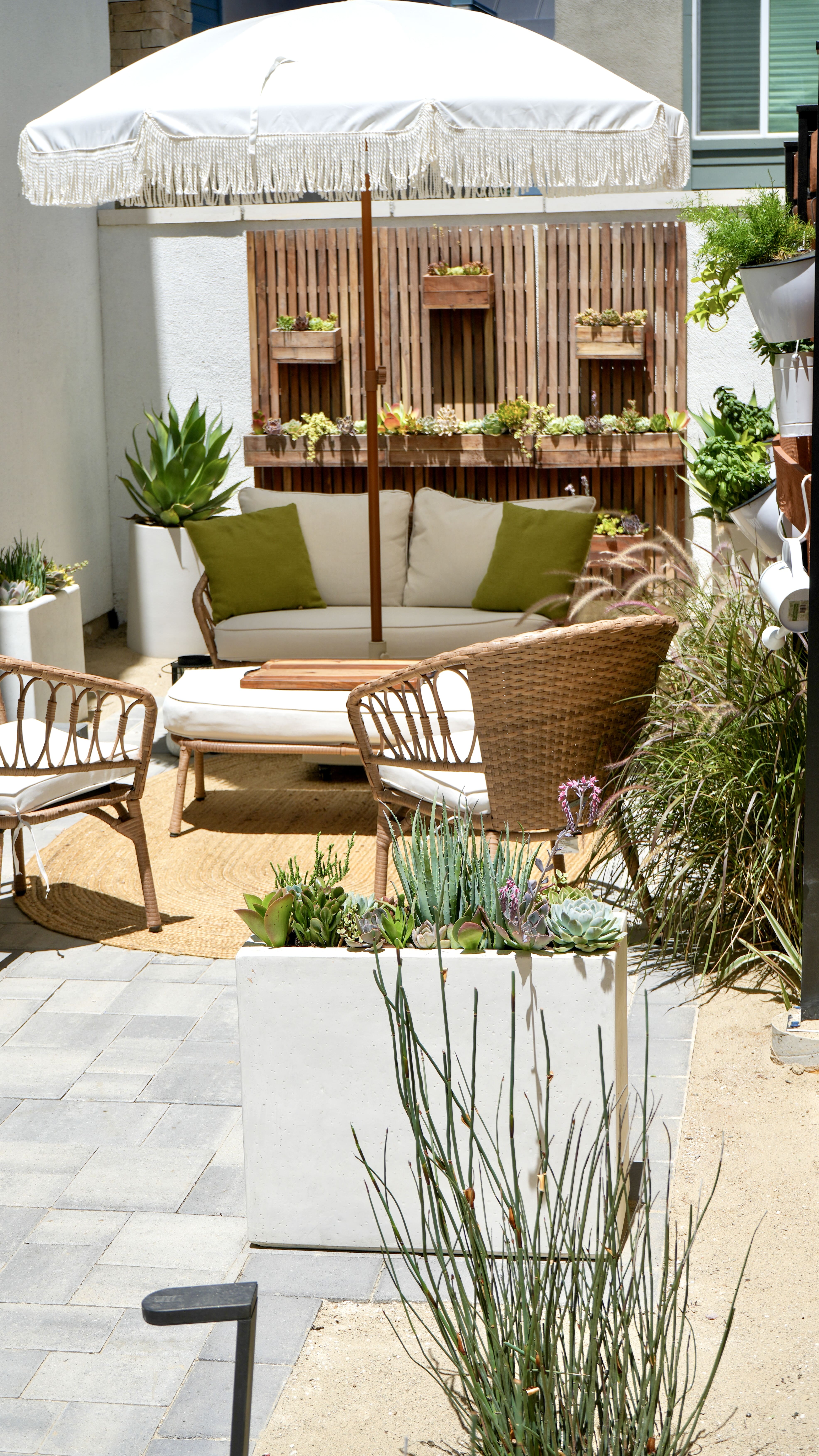 Shot of colorful side patio. Showing a love seat couch, table, plus two chair and an umbrella. Also shown is a large succulent wall planter and herb garden vertical wall with white pots.