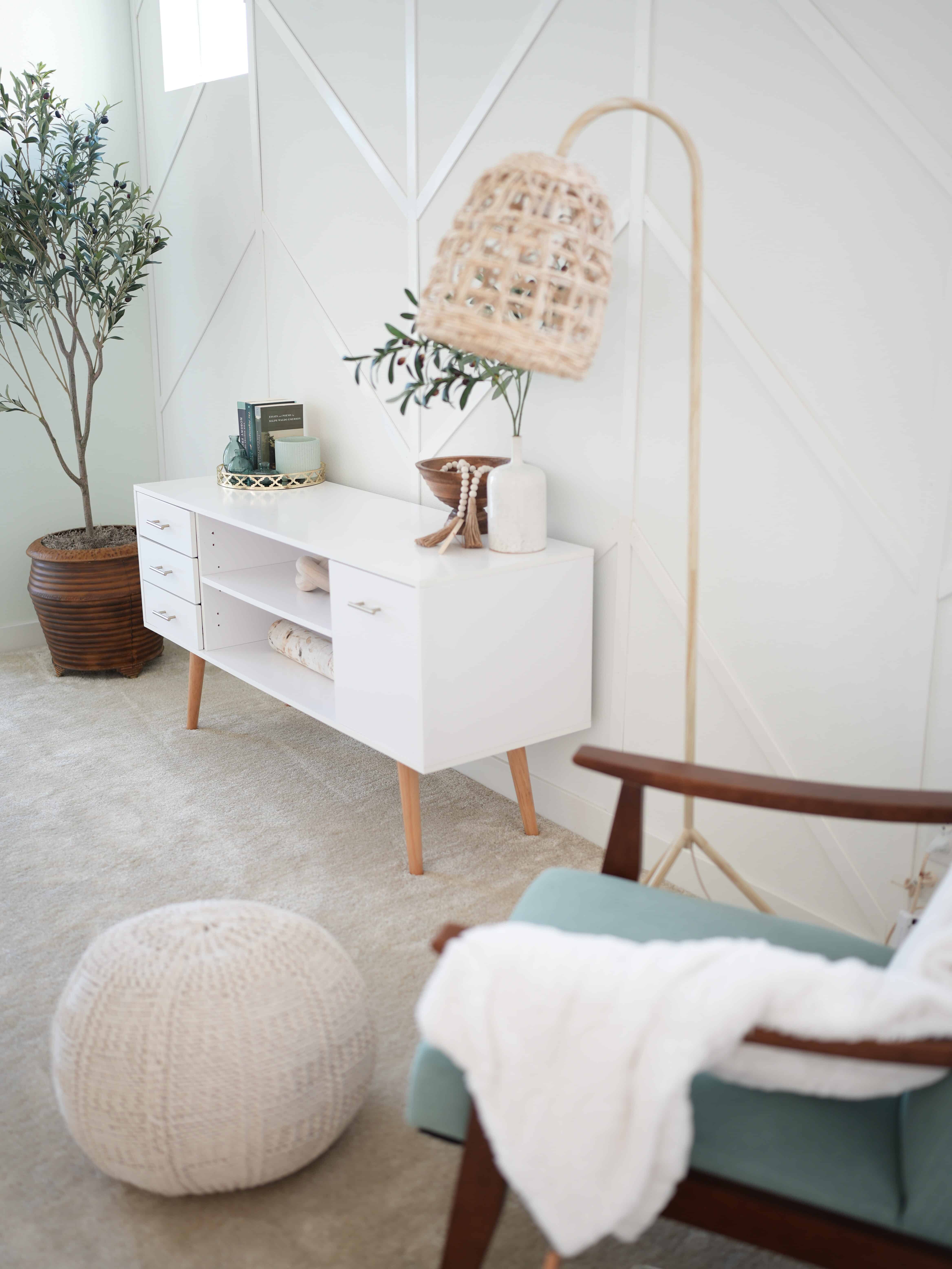 Corner shot of a reading nook room. Long end table with decor, tall olive tree in brown planter in the corner, with a teal colored chair on the opposite side, with a white blanket thrown over it and a round, beige foot rest.