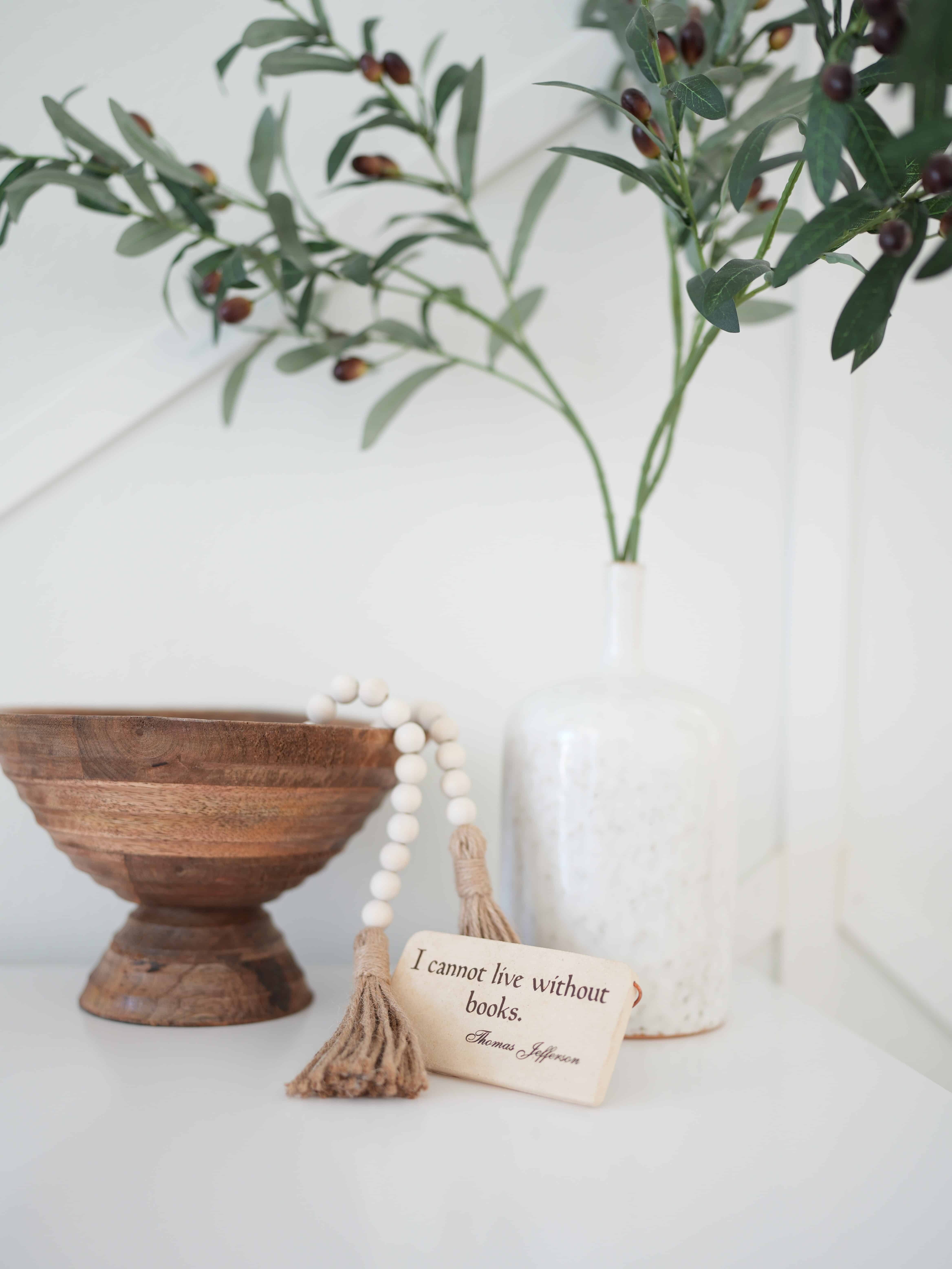Brown decorative bowl with decorative beaded string spilling out of it. Next to a small beige bookish sign that reads "I cannot live without books" - Thomas Jefferson, in front of a tall white vase with faux green stems, sitting on top of a white table against a white wall.