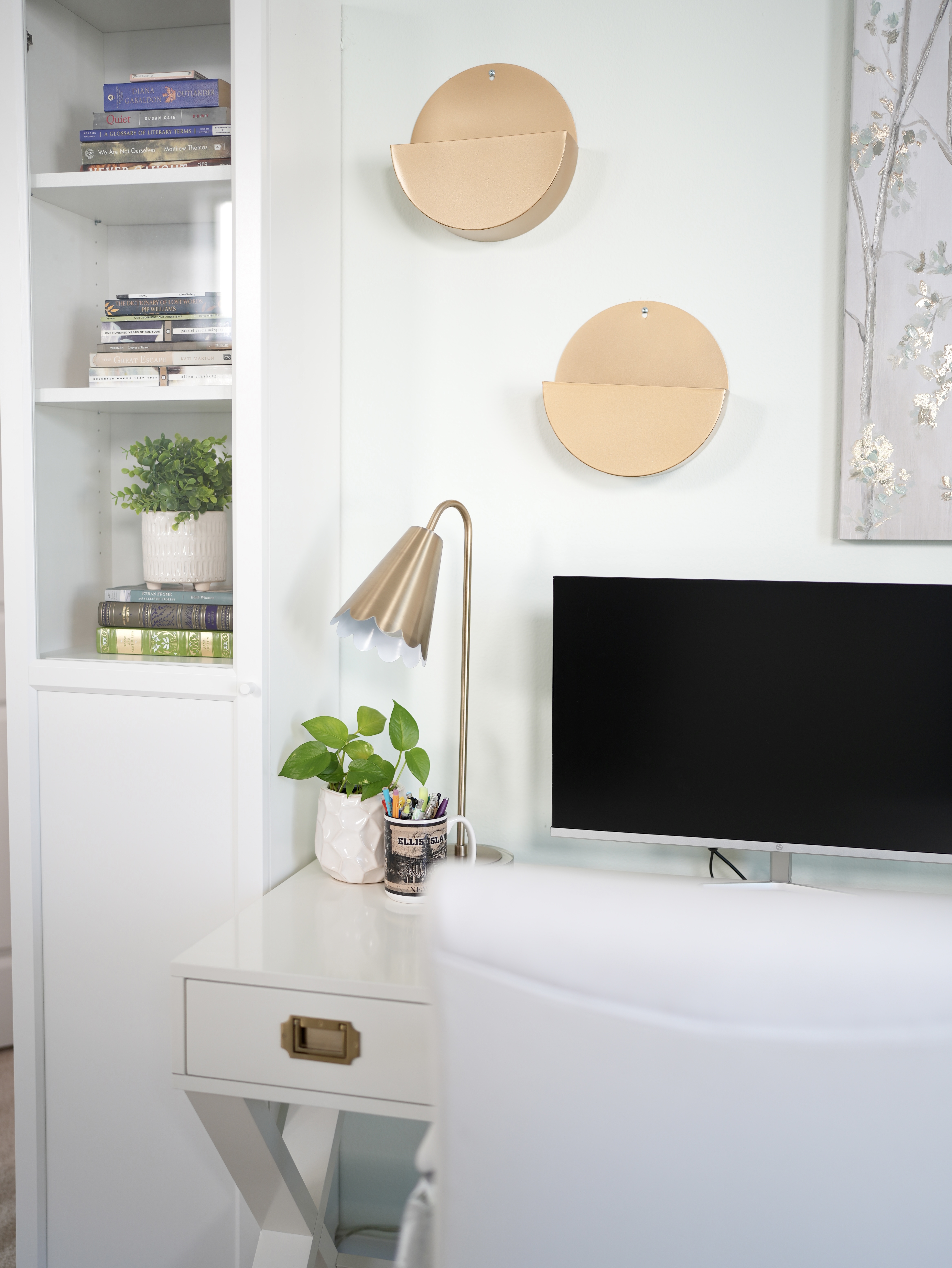 A white desk next to a white bookshelf with glass window showing several paperback books and faux plant. Desk contains a black computer screen, gold tall reading light, Ellis Island coffee mug with pens inside, and a small white planter with a green pothos plant. Above it on the wall, a golden round decorative accent and part of a teal and golden square wall art piece.