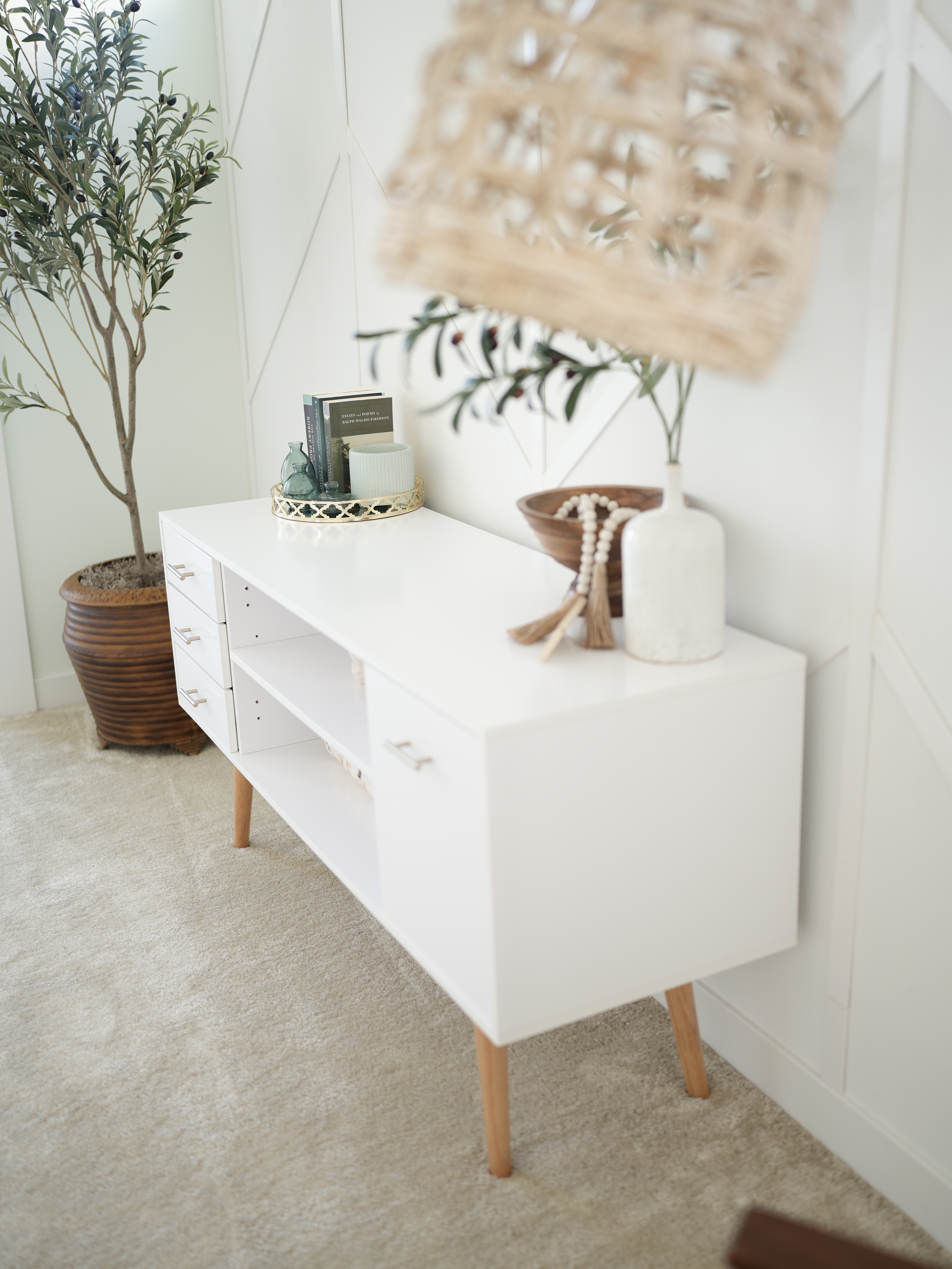 large faux olive tree in brown pot standing in corner of the room. White large console table with home decor and paperback books displayed. White decorative wall. Top part of lighting fixture displayed in top corner on the shot.