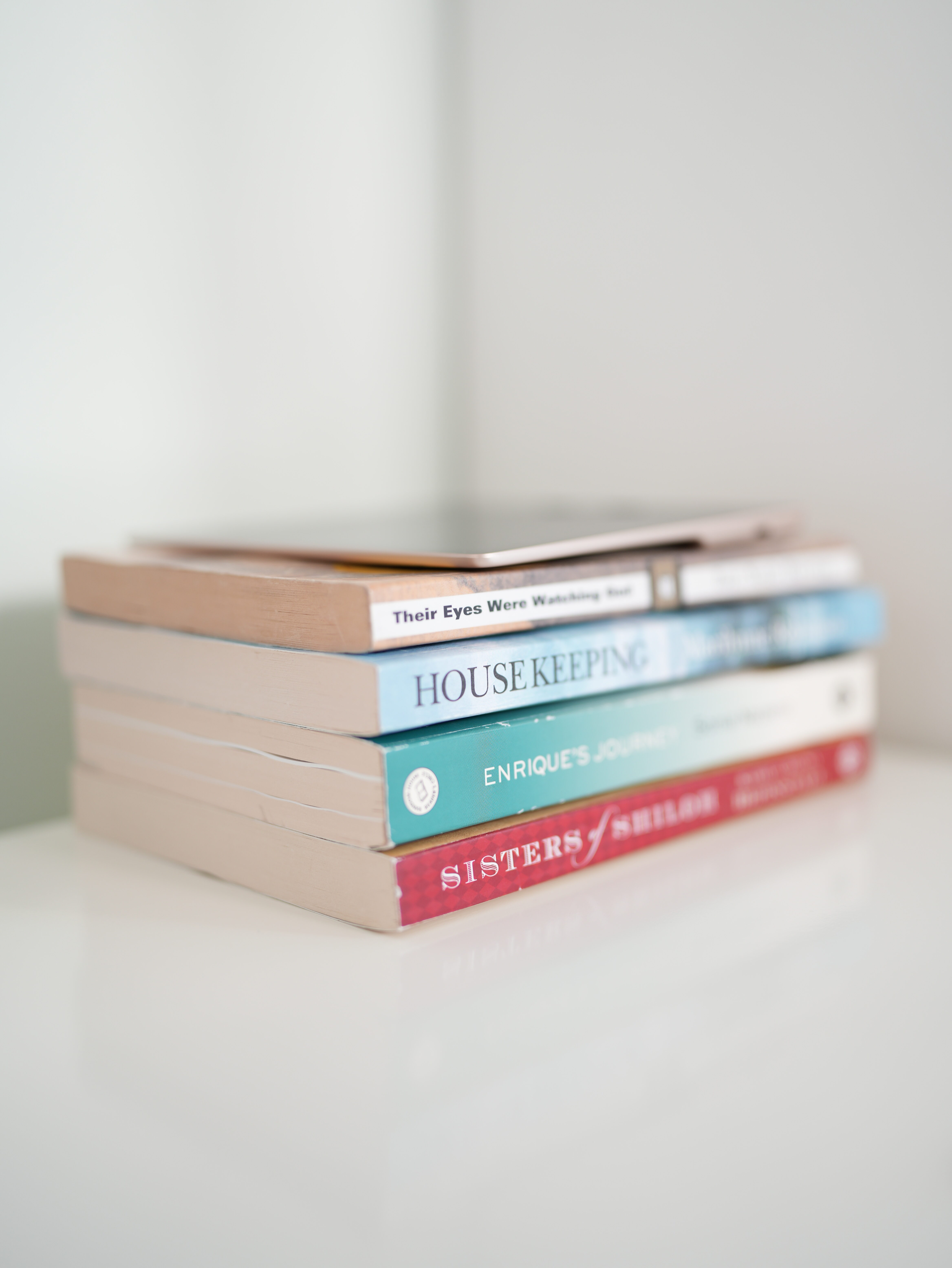 Photo of four colorful paperback books laying horizontally on top of each other, with a thin Amazon Kindle on top, on white background.