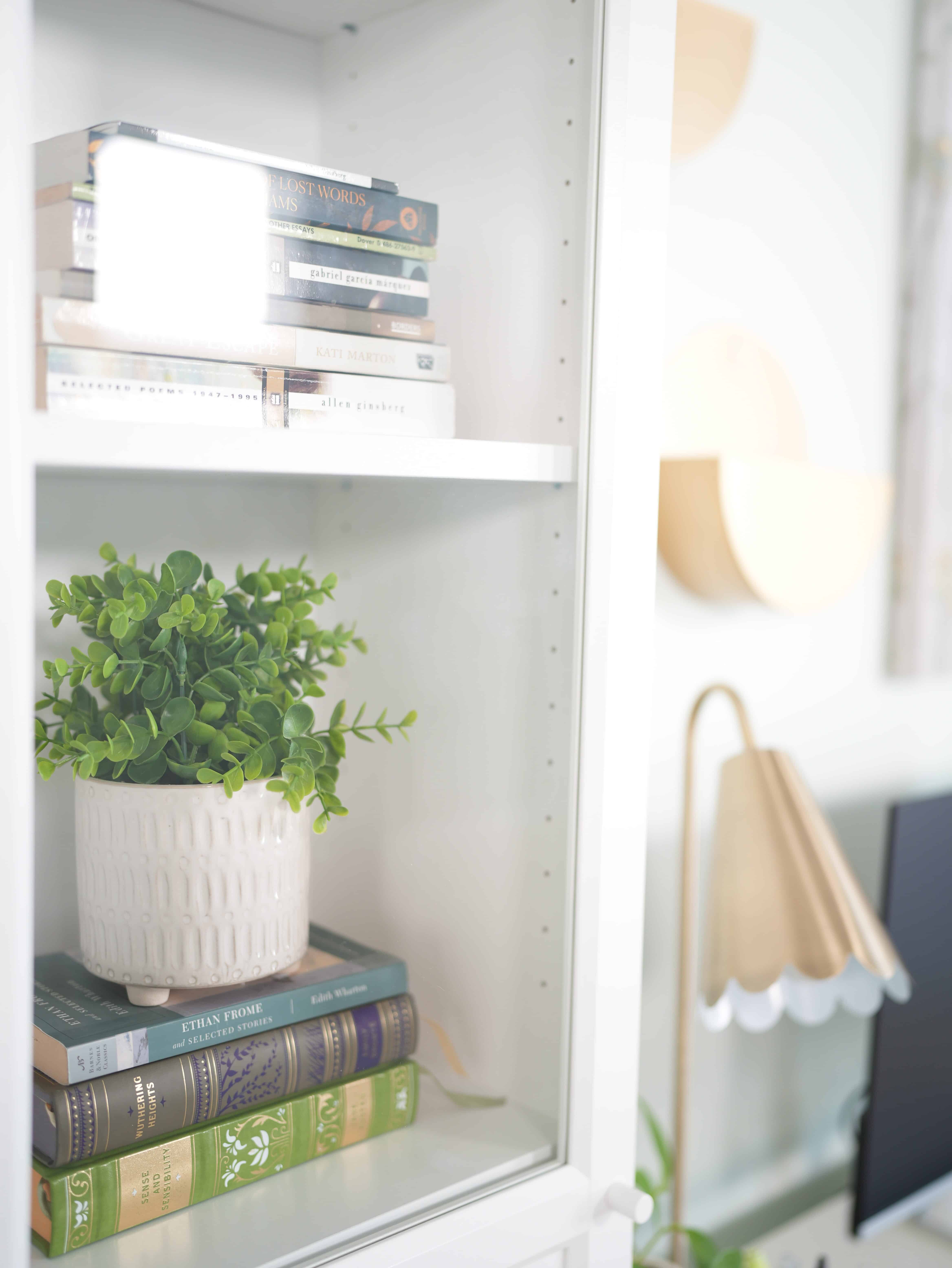 Glass encased books, inside a tall and narrow bookshelf. Paperback books laying on top of each other. In the distance shows a golden desk lamp, black computer screen, and white wall with golden wall art accents.