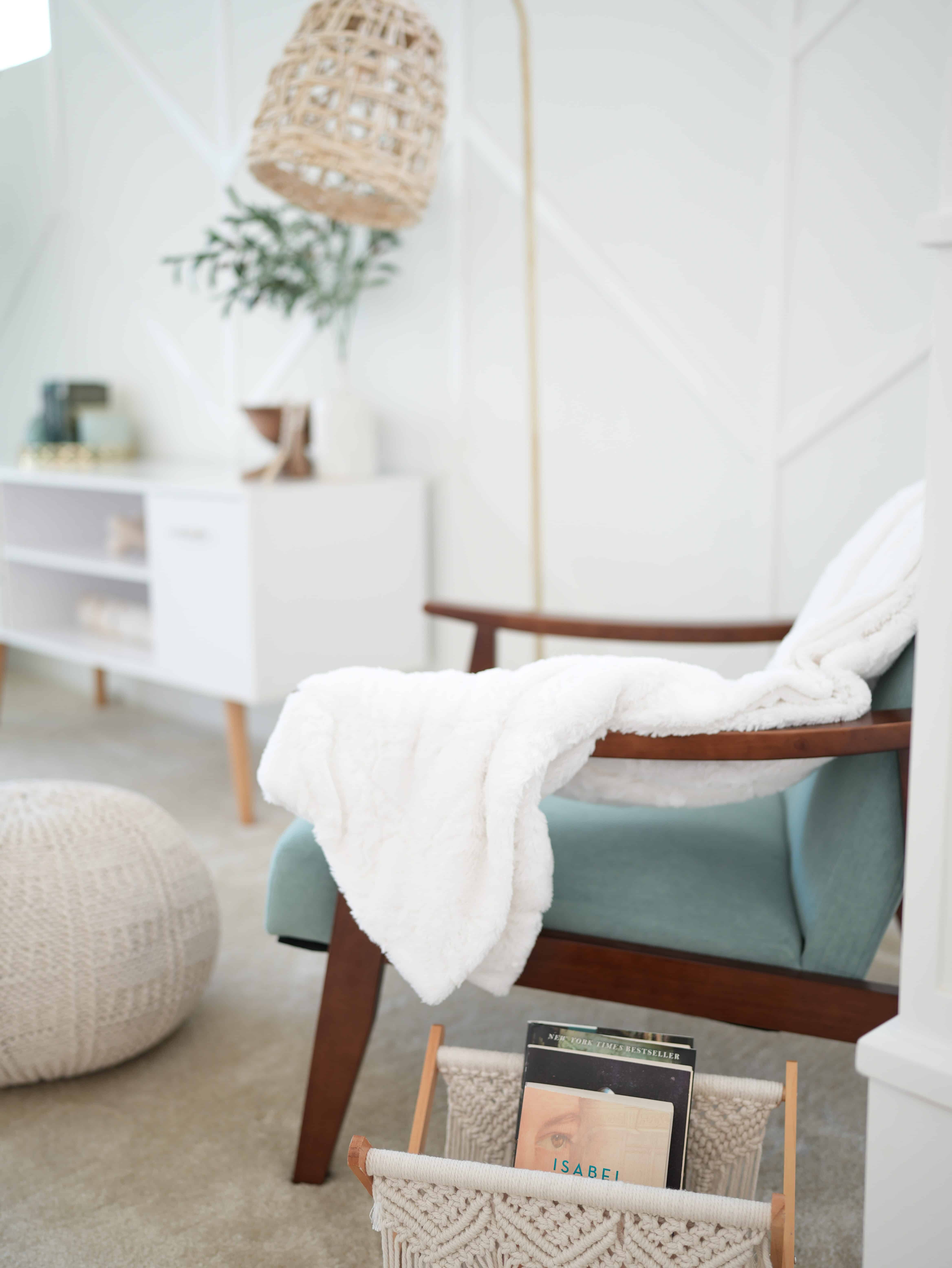 Corner shot of a reading nook. Showing a teal chair with a white throw thrown over it, a beige foot stool, a tall light fixture, and a white end table with decor, against a white wall.