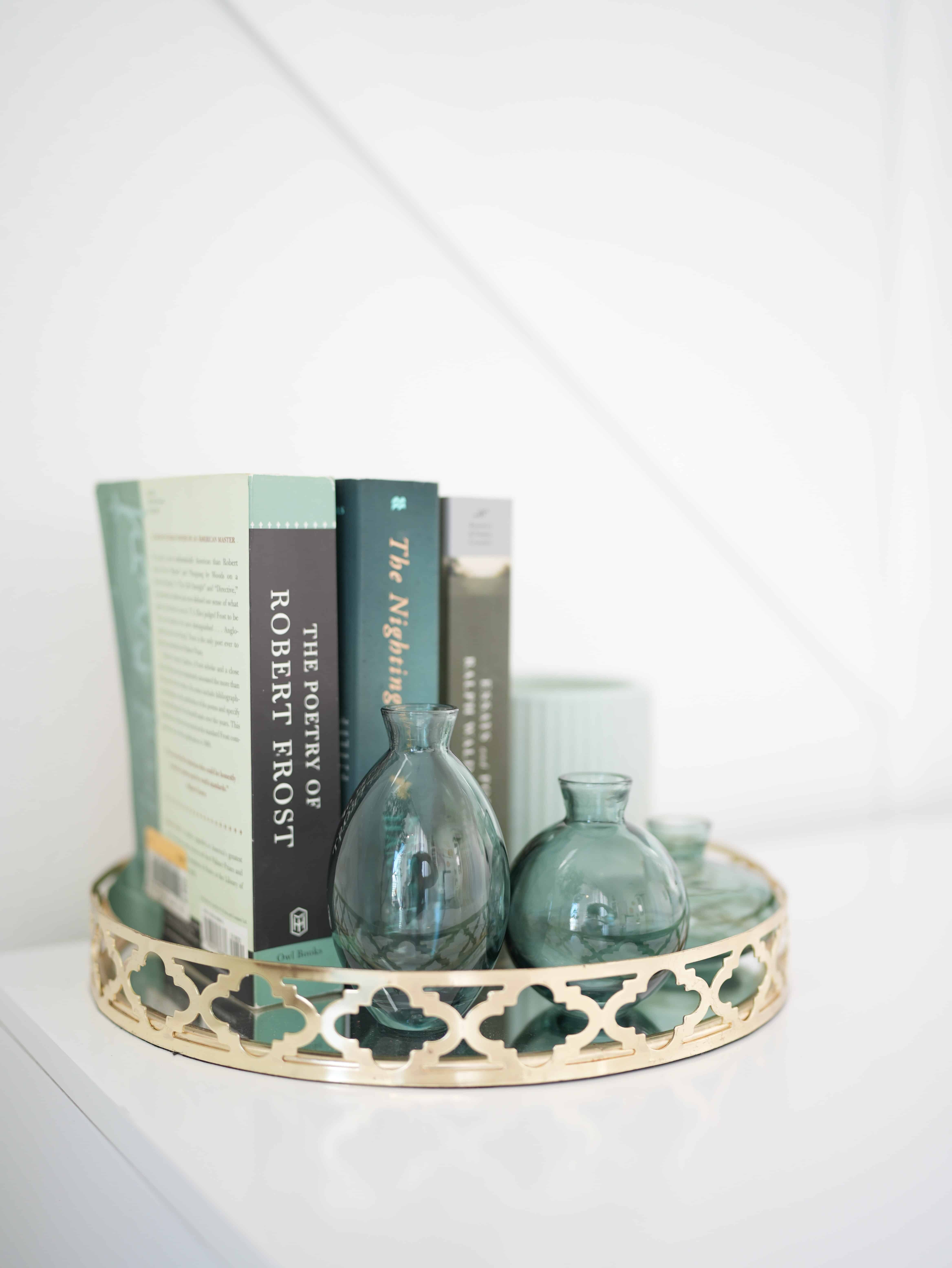 Round and shallow golden tray with three small glass vases inside, along with three thick paperback books in shades of blue and green, and a teal candle.