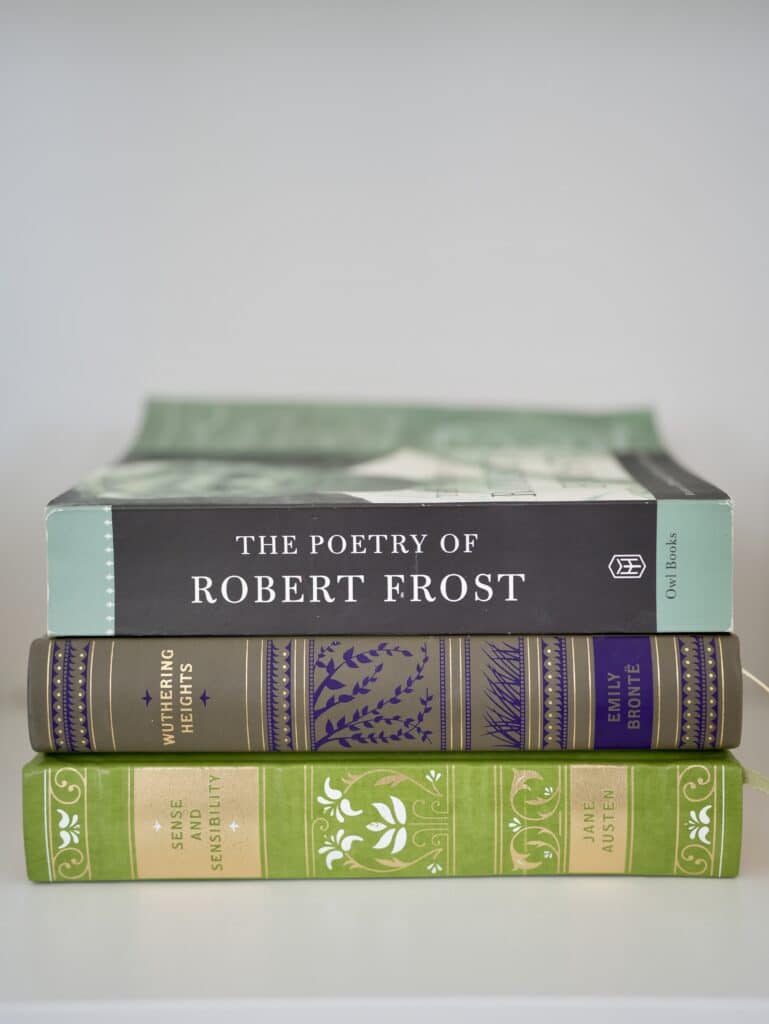 Three thick paperback novel books laying on top of each other in white bookcase. Green covered book by Jane Austen, Purple and dark grey covered book by Emily Bronte, teal and black covered book by Robert Frost.