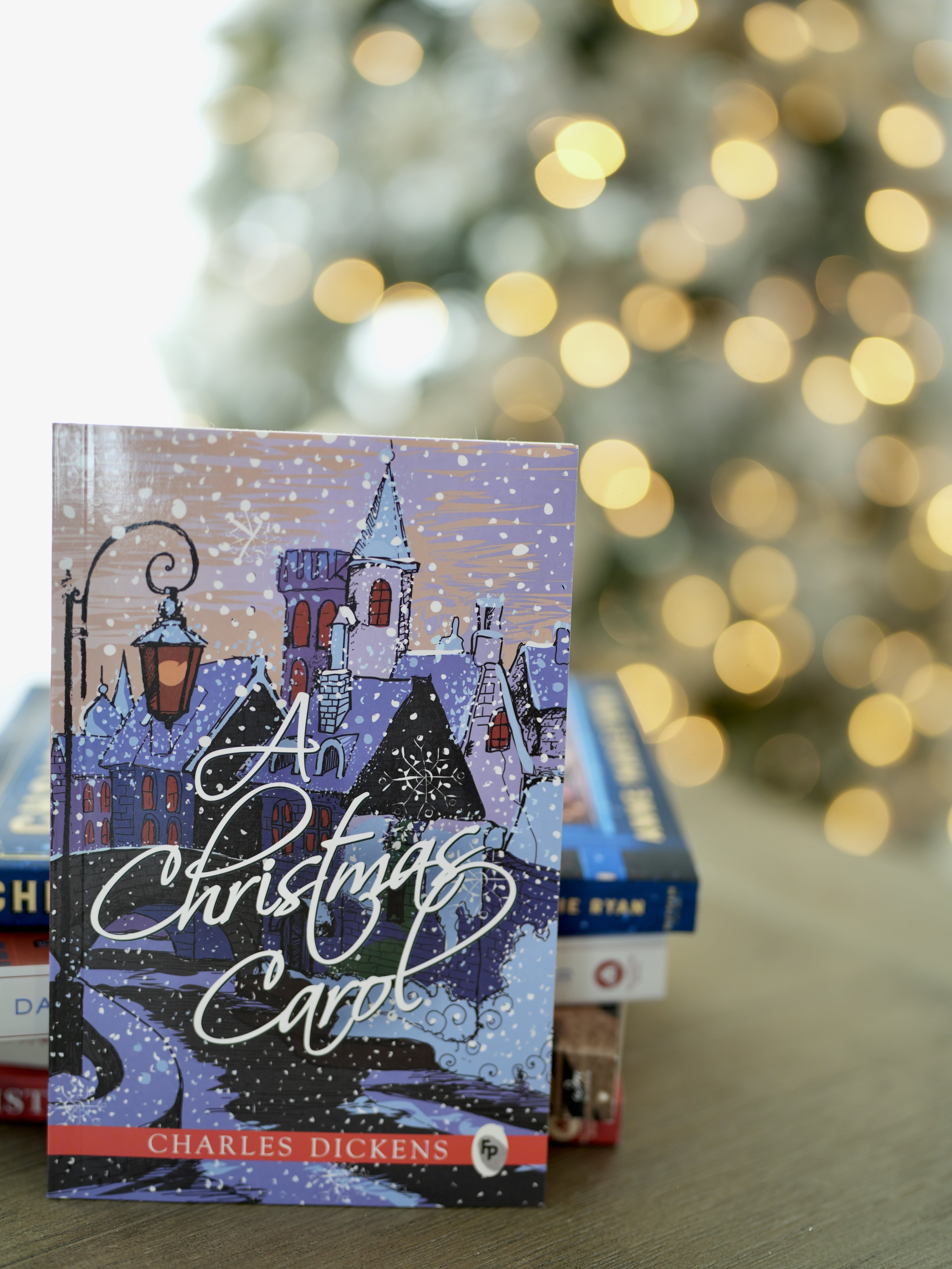Photo of a book, titled A Christmas Carol, by Charles Dickens. Book is standing upright behind a stack of books, and on top of a brown table. Also sits in front of a lit up Christmas tree.