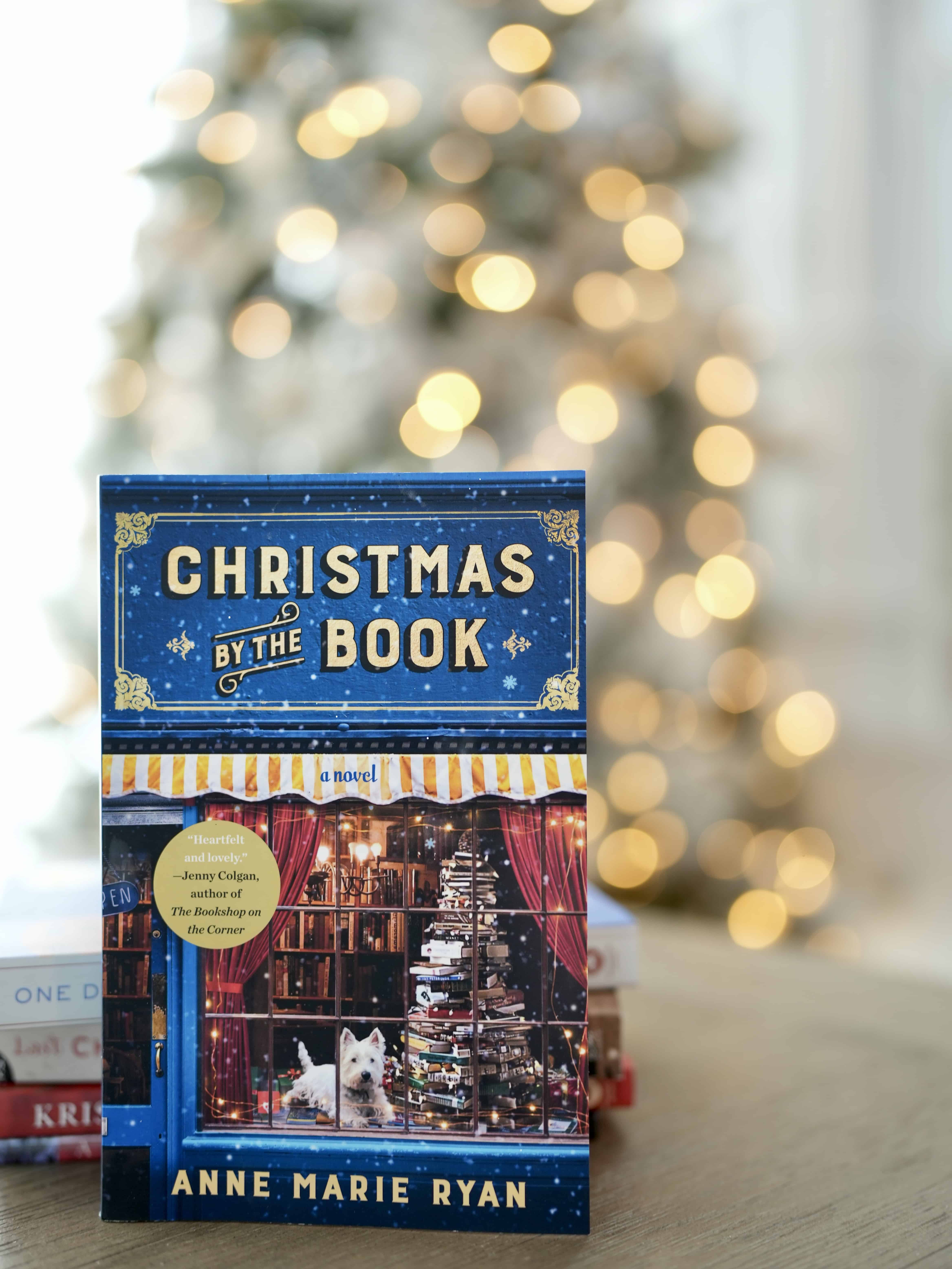 Photo of a book, titled Christmas by the Book, by Anne Marie Ryan. Book is standing upright behind a stack of books, and on top of a brown table. Also sits in front of a lit up Christmas tree.