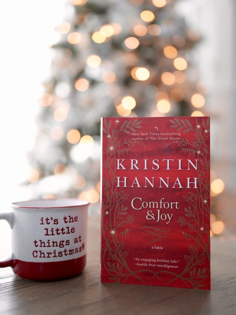 Comfort & Joy book by Kristin Hannah, sits next to a small Christmas-themed coffee cup, in front of a lit-up Christmas tree.