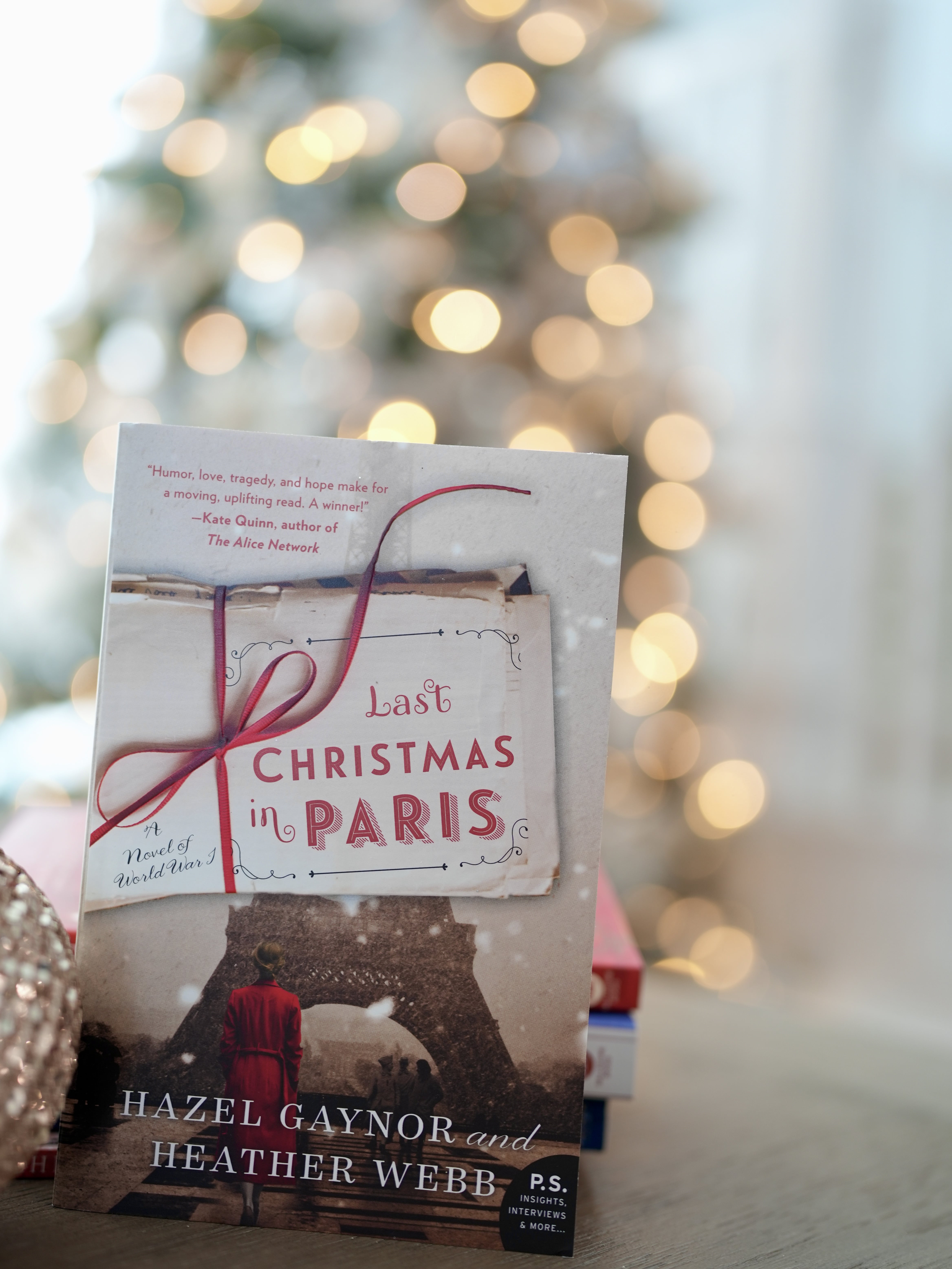 A book titled The Last Christmas in Paris, stands behind a stack of books on a brown table. A lit up Christmas tree is shown in the background.