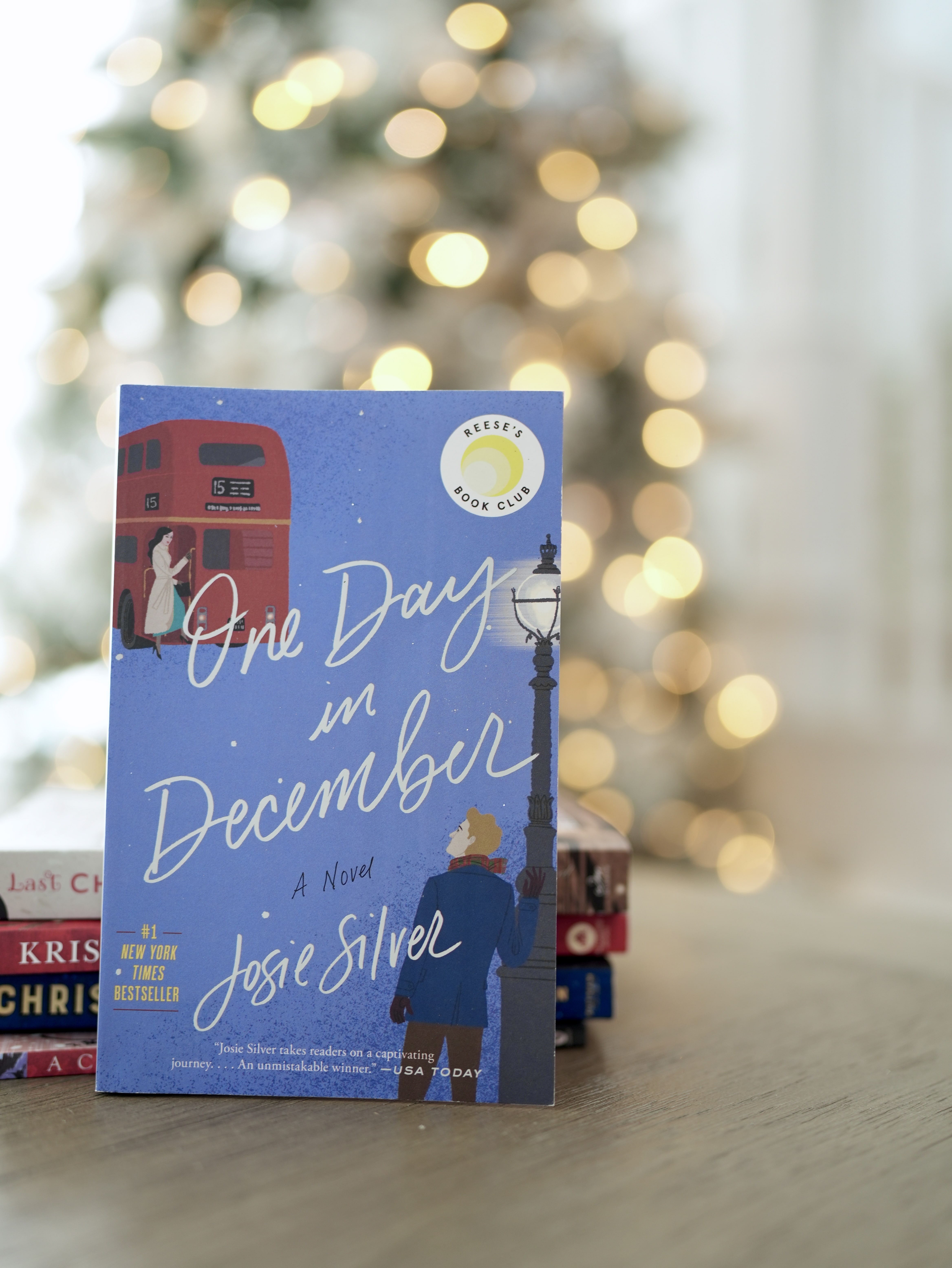 The book, titled One Day in December, by Josie Silver, stands behind a stack of books on a brown table. A lit up Christmas tree is in the background.