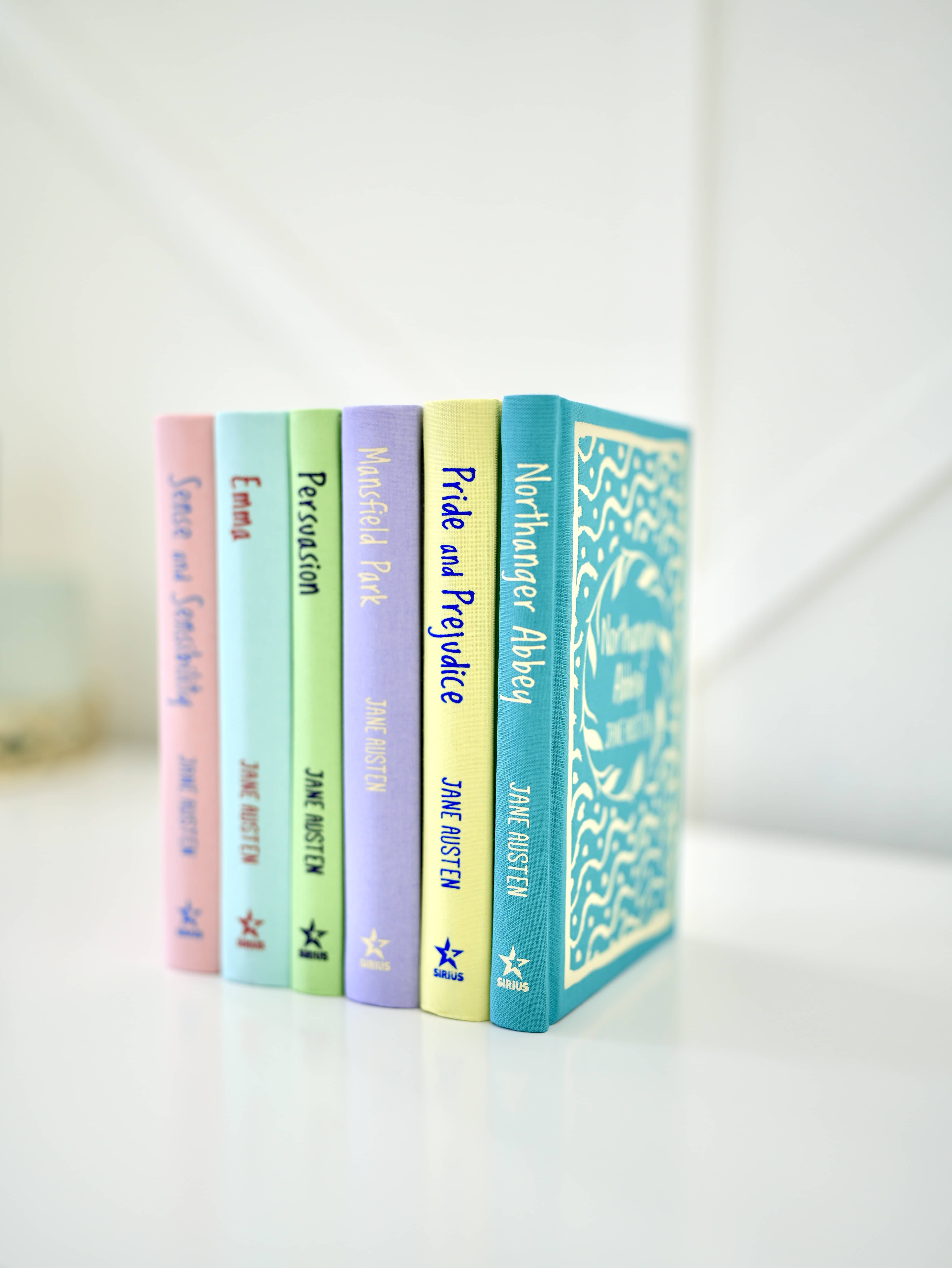 Jane Austen hardcover books standing on white table in front of white wall. Multi-colored books.