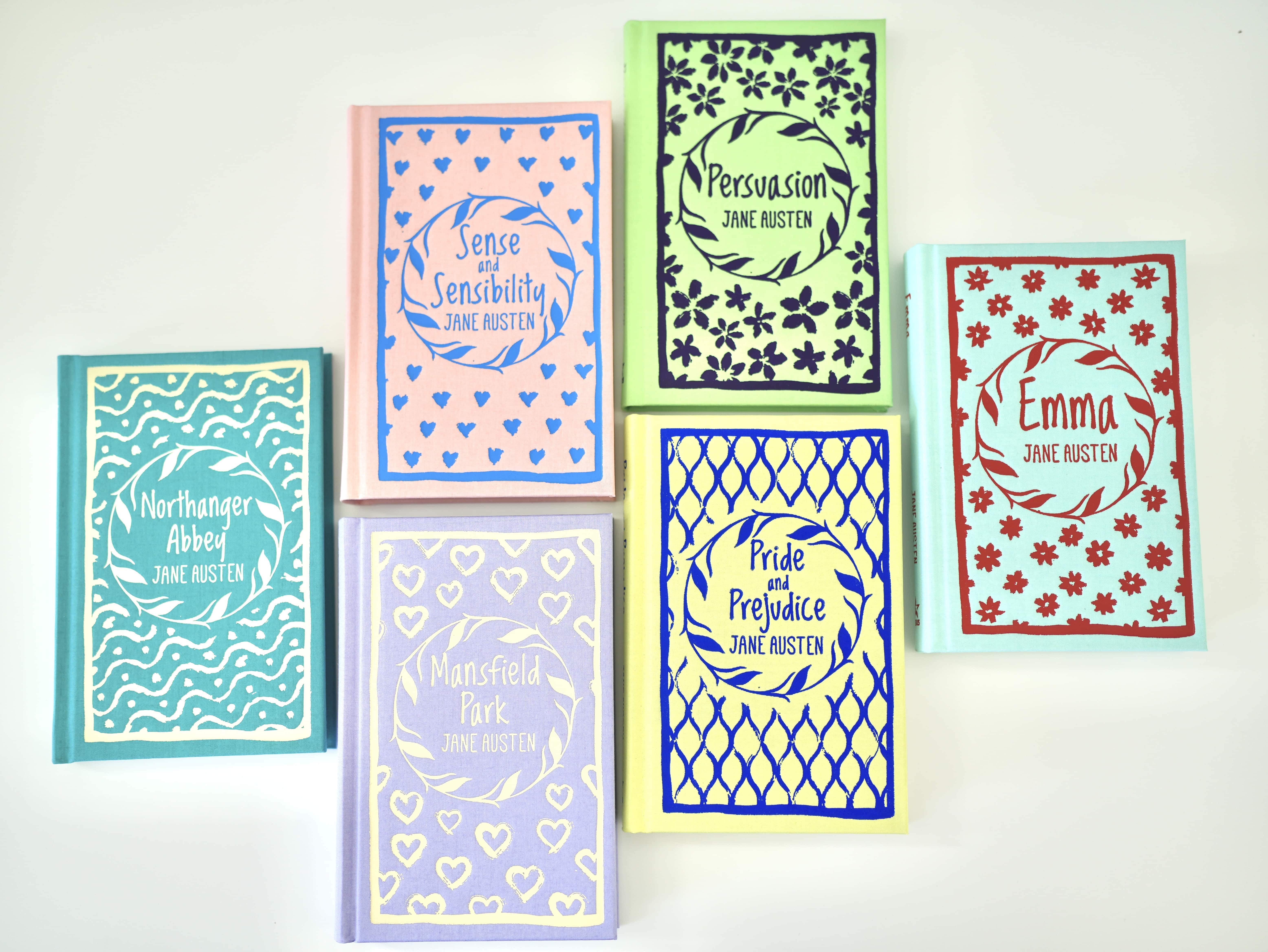 Photo of six Jane Austen novels laying on white background. Northanger Abbey with teal cover, Sense and Sensibility with Pink Cover, Mansfield Park with lavender cover, Persuasion with lime cover, Pride and Prejudice with yellow cover, Emma with light blue cover.