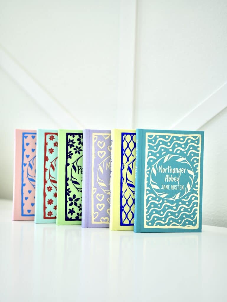 Jane Austen hardcover books with individual covers in pink, teal, green, lavender, yellow, and blue. Standing on white table with white background.