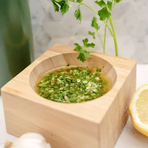 Chimichurri sauce sitting in wooden container, next to parsley plant, garlic bulb, sliced lemon and a green bottle of olive oil.