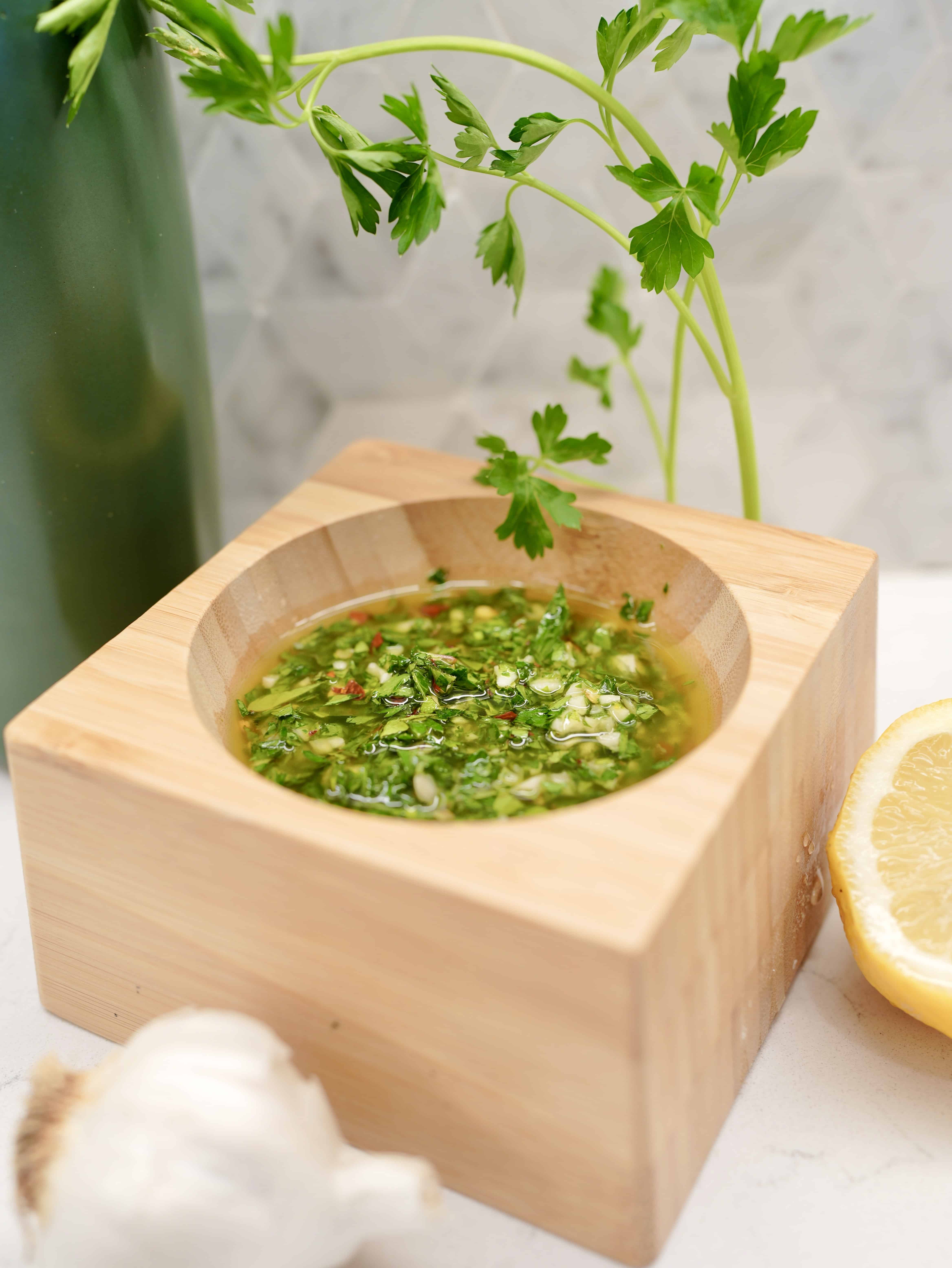 Chimichurri sauce sitting in wooden container, next to parsley plant, garlic bulb, sliced lemon and a green bottle of olive oil.