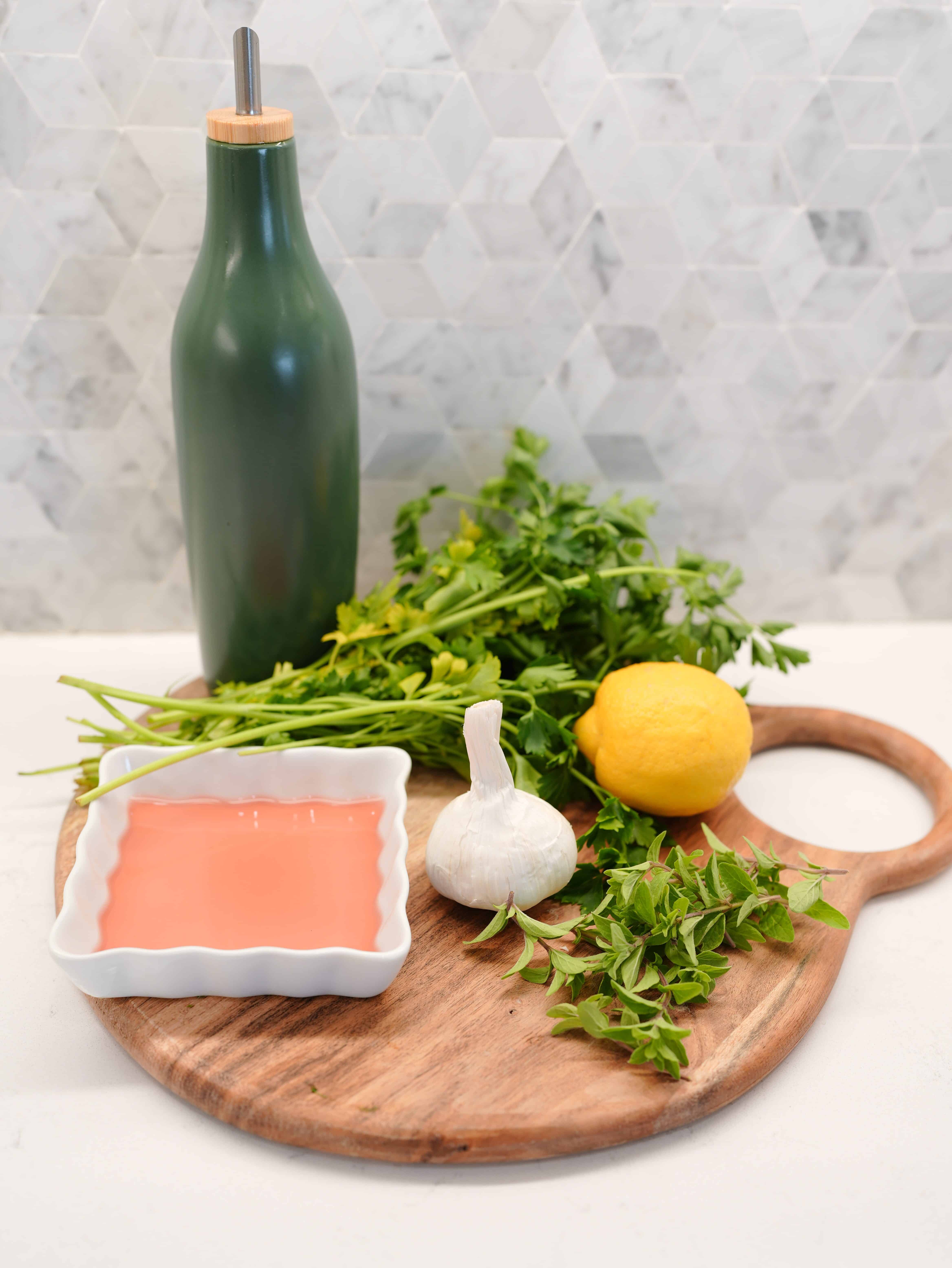 Ingredients of Chimichurri sauce: Olive oil, red wine vinegar, lemon, garlic bulb, parsley stems, on a wooden cutting board