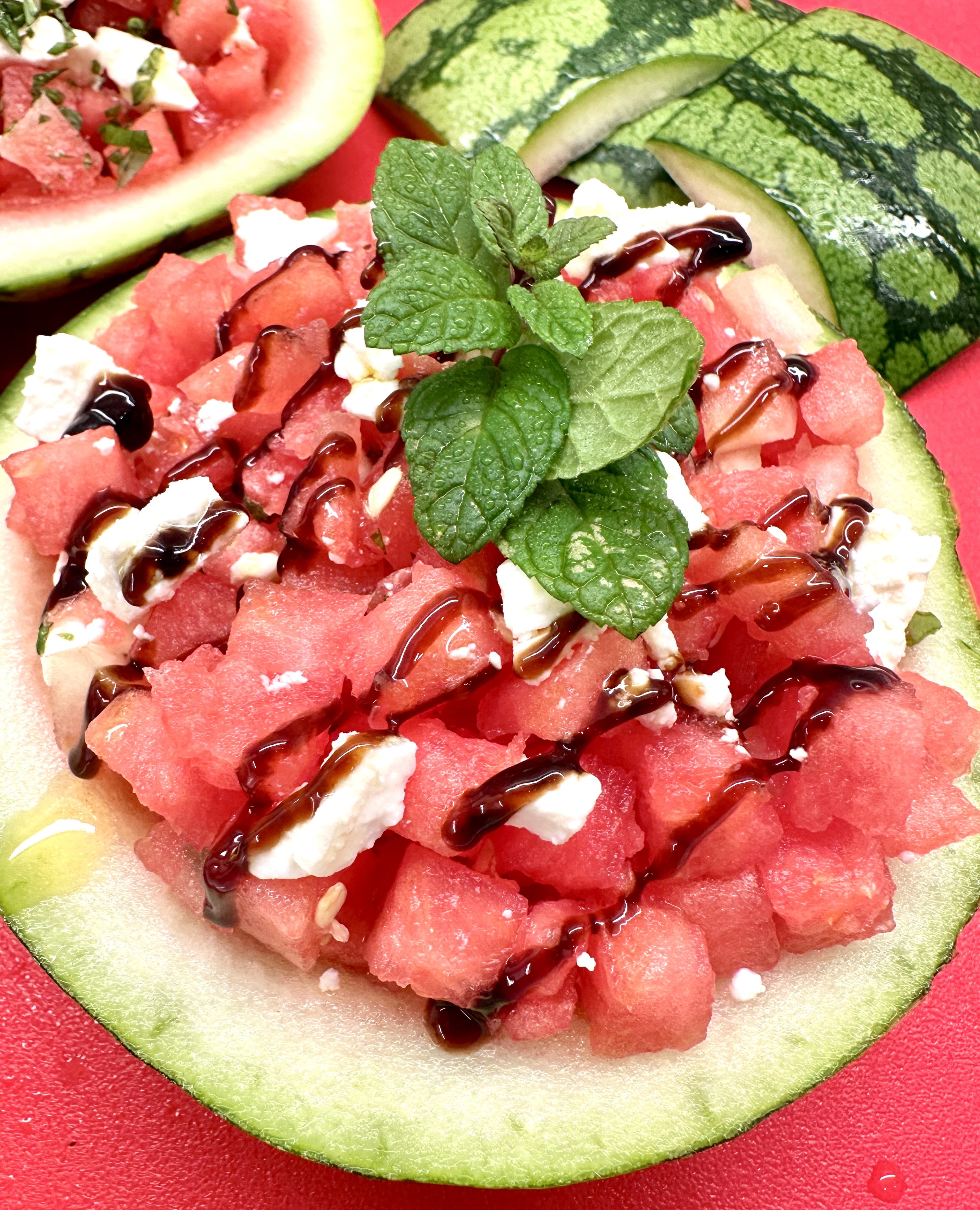 Watermelon salad with crumbled feat cheese and fresh mint leaves, sitting inside watermelon husk on red cutting board.