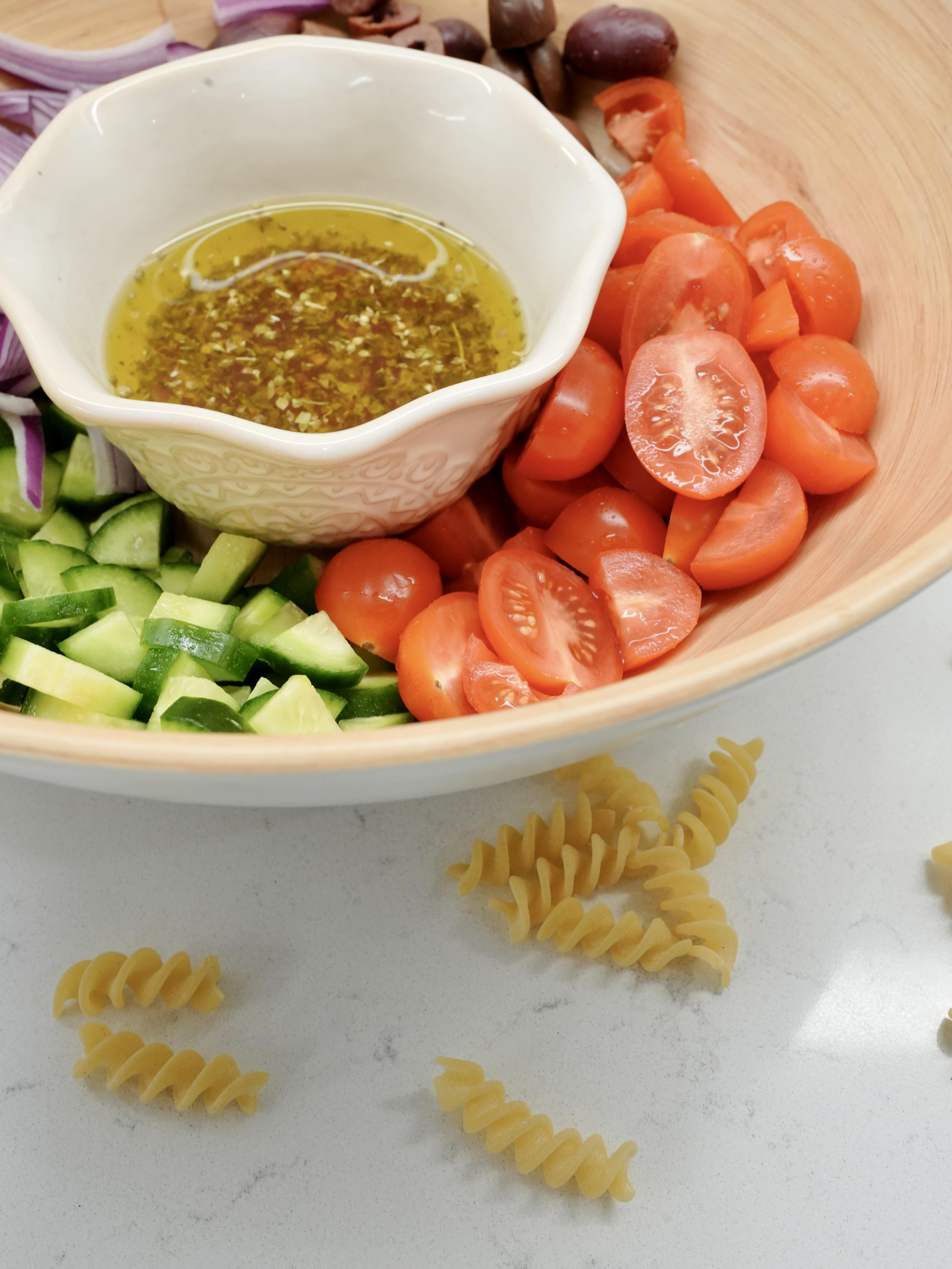 Ingredients for Greek Pasta Salad: Chopped cucumbers, sliced cherry tomatoes, sliced olives, a bowl of vinaigrette, served in a large serving bowl