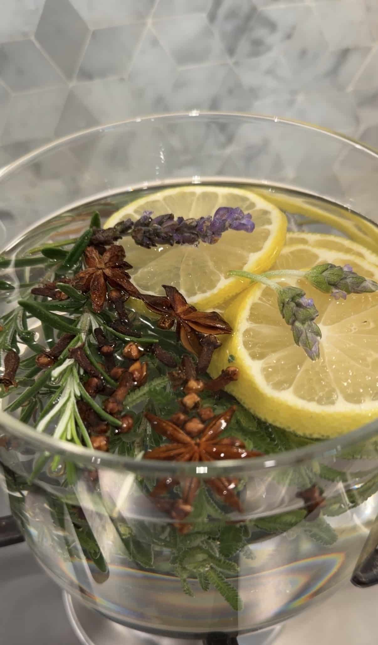 Sliced lemon, star anise, cloves, and lavender flowers simmering in glass pot over the stove